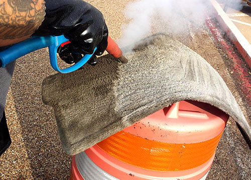 Person steam cleaning a carpeted car floor mat